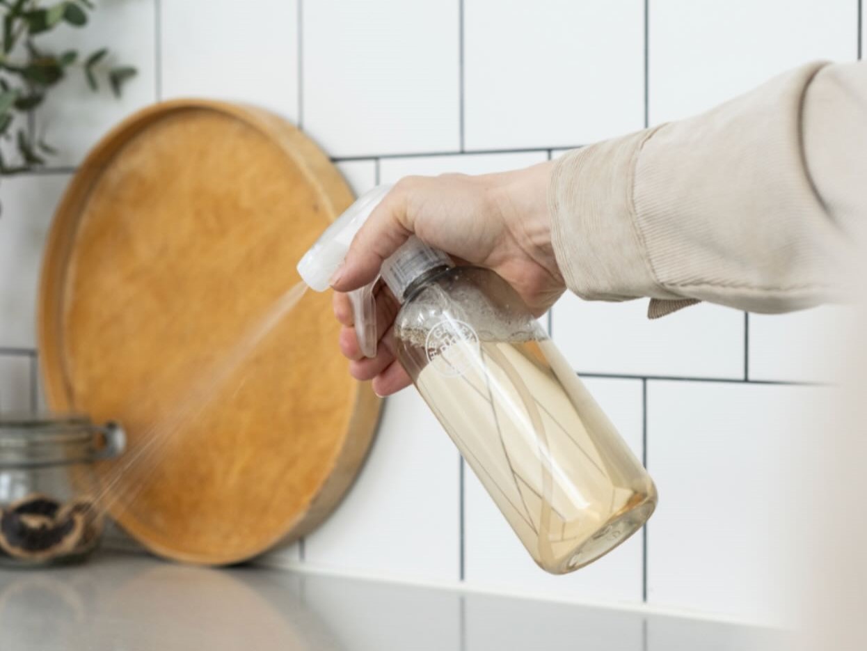 Hand spraying a reusable glass cleaner bottle onto a tiled kitchen backsplash. The Green Lab Co